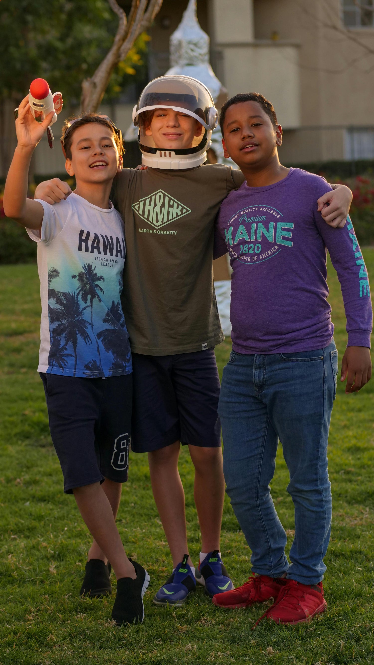 Three children posing together outdoors, with one wearing a space helmet.